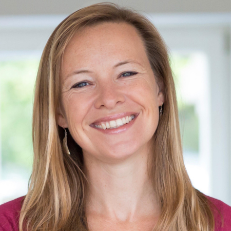 Miranda is standing inside in front of a window. She is smiling at the camera and is wearing red t-shirt.